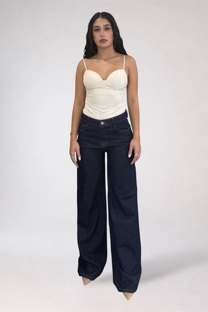 A woman modeling a cream-colored ribbed camisole top with spaghetti straps and dark wide-leg denim jeans against a plain light gray studio background.