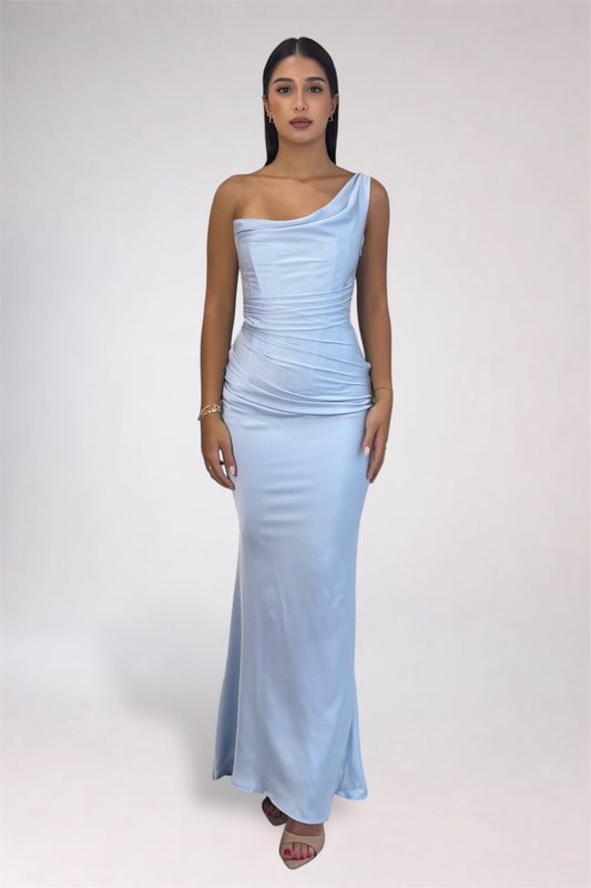 A woman modeling a light blue one-shoulder maxi dress with a ruched bodice and flared skirt, standing against a plain light gray studio background.