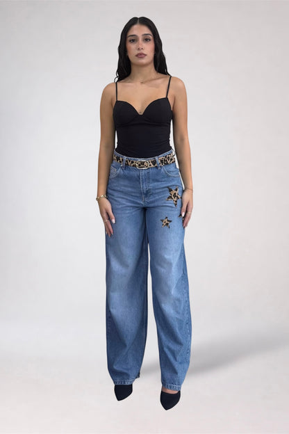 A model stands against a light gray studio background wearing a black spaghetti-strap camisole top, wide-leg blue jeans with embroidered star details on the thighs, and a patterned belt.