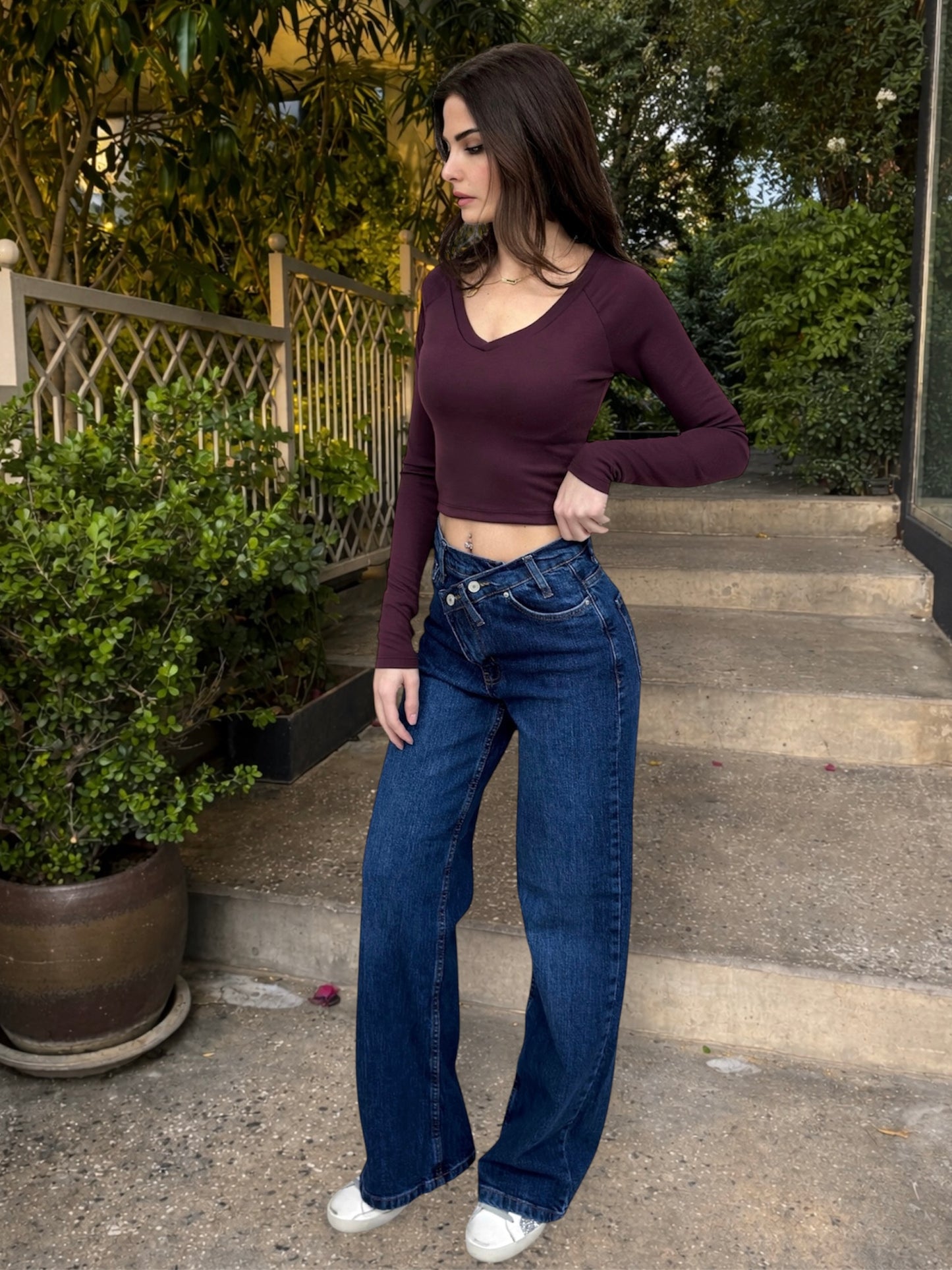 Woman wearing a burgundy long-sleeve top and blue jeans standing on steps outdoors.