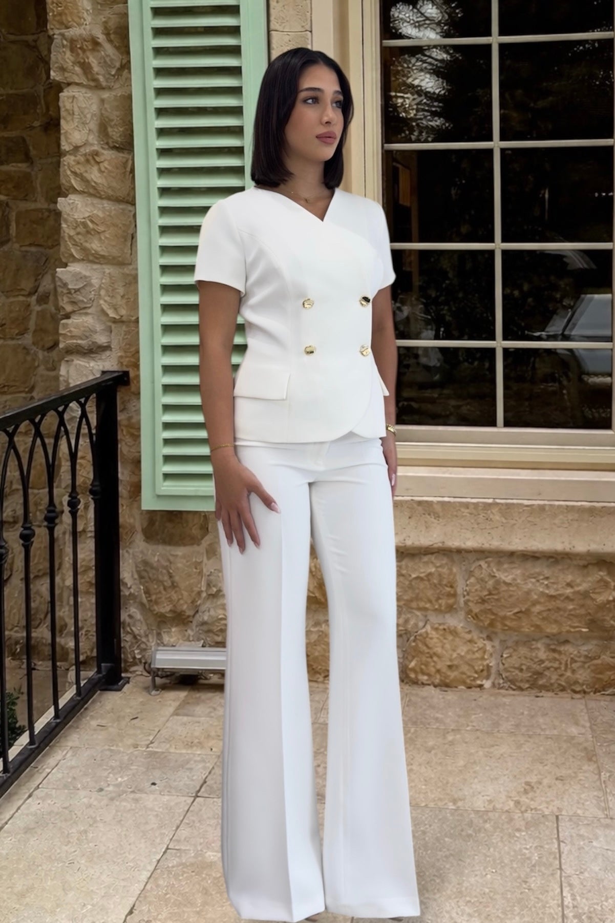 Woman in a white outfit standing in front of a stone building with green shutters.