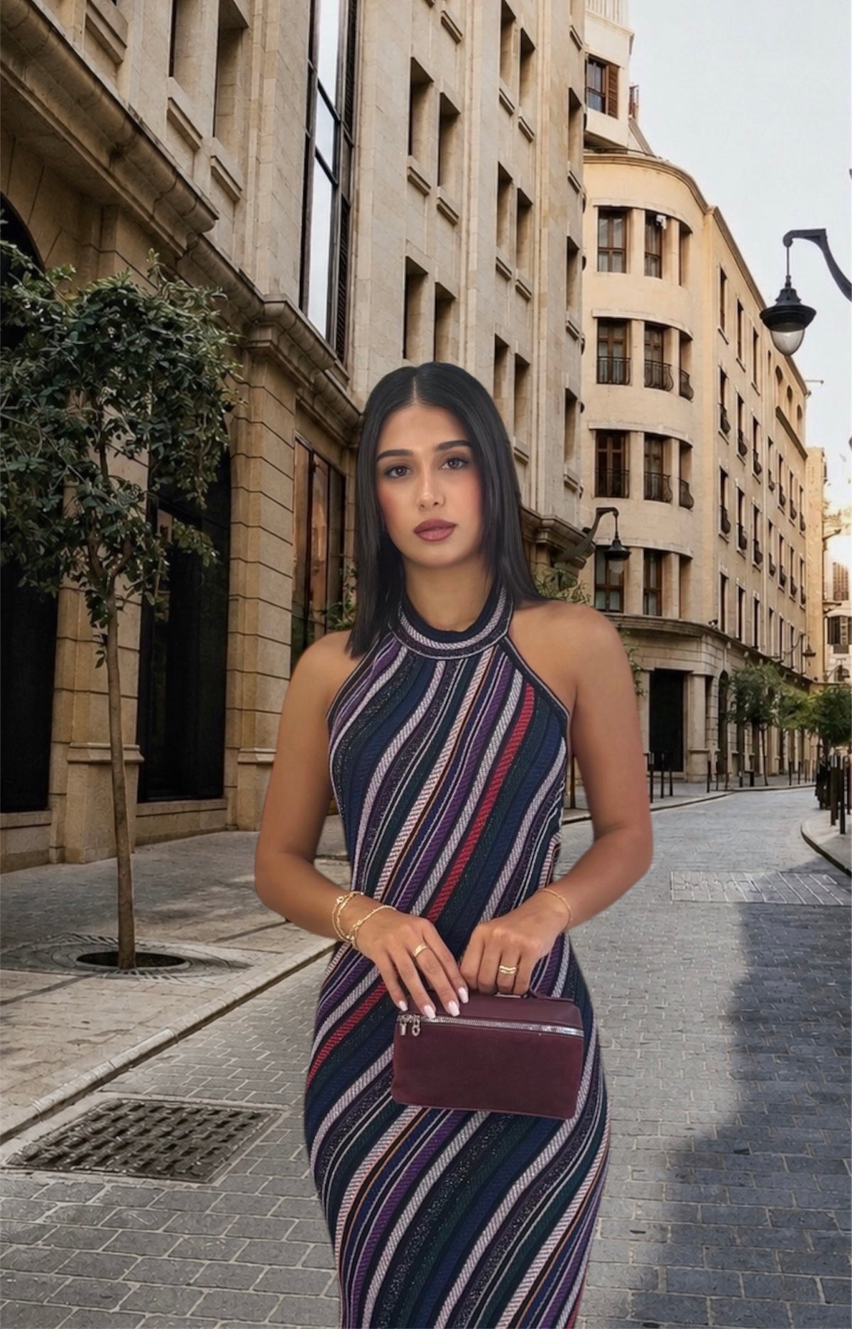 A woman stands on a city street wearing a sleeveless, high-neck, bodycon dress with vertical navy, red, and white stripes. She is holding a small burgundy clutch bag.