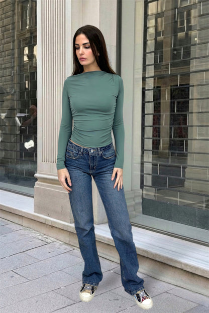 Woman wearing a green long-sleeve top and blue jeans standing on a city street.