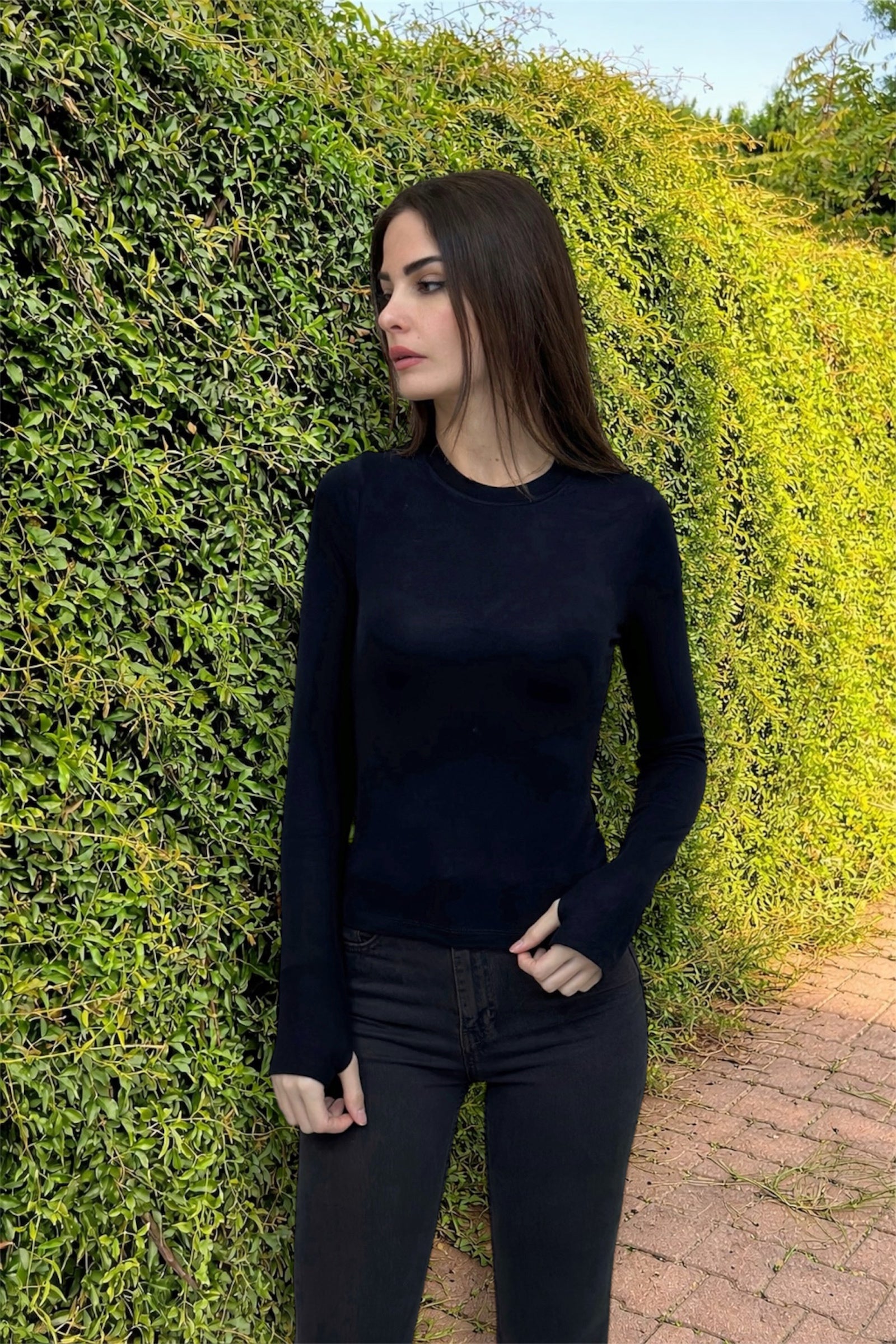 Woman wearing a black long-sleeve top and dark pants standing against a green hedge.