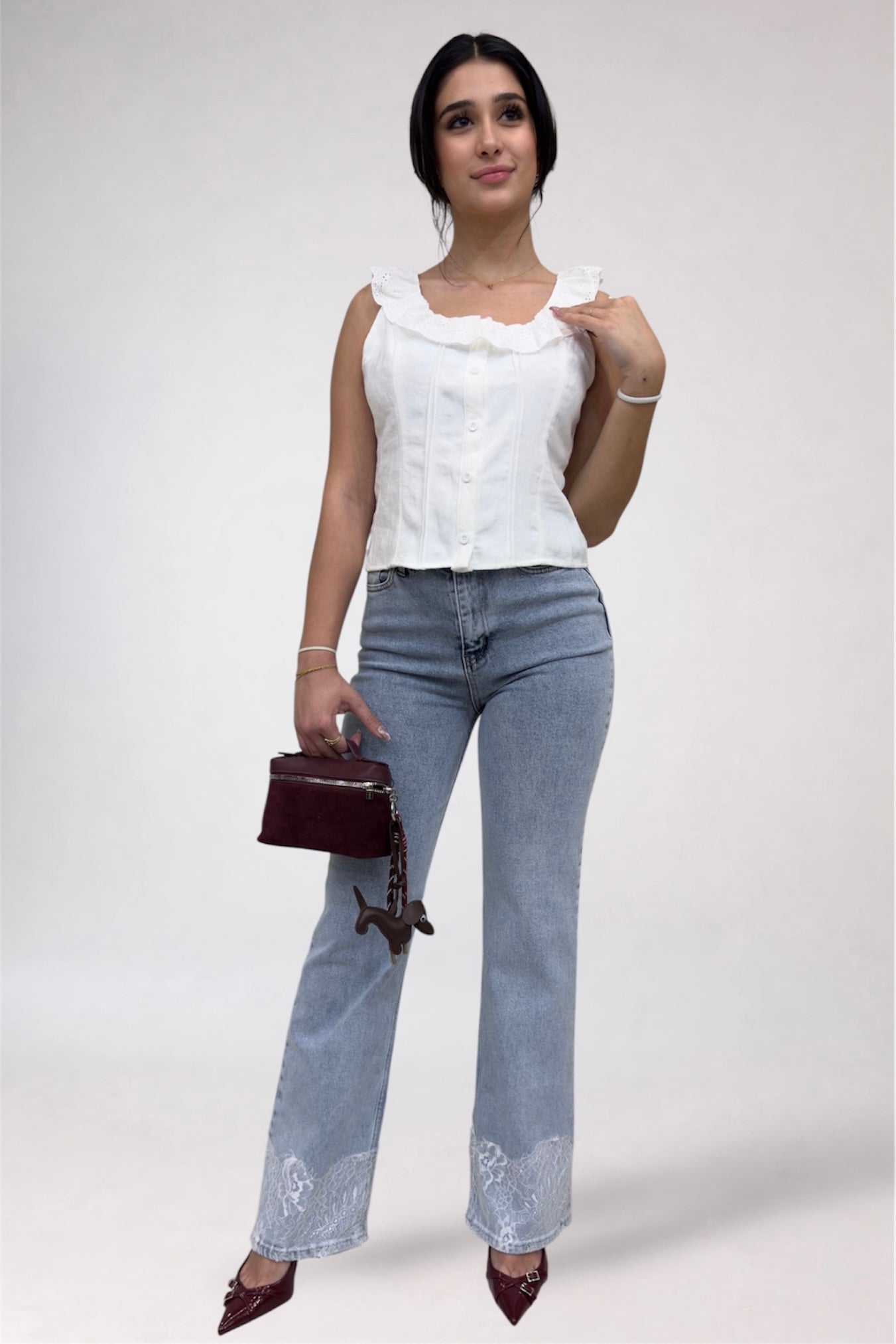 Woman wearing a white sleeveless top and light blue jeans with a small handbag on a plain background