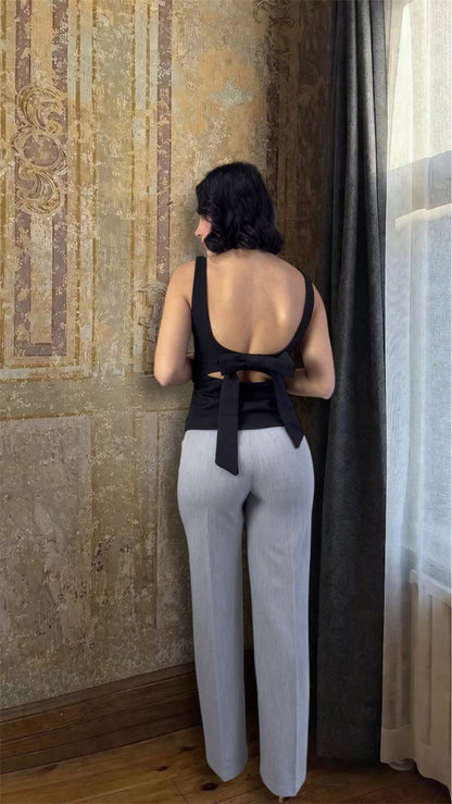 Woman wearing a black top and light gray pants standing in a room with patterned wallpaper and curtains.