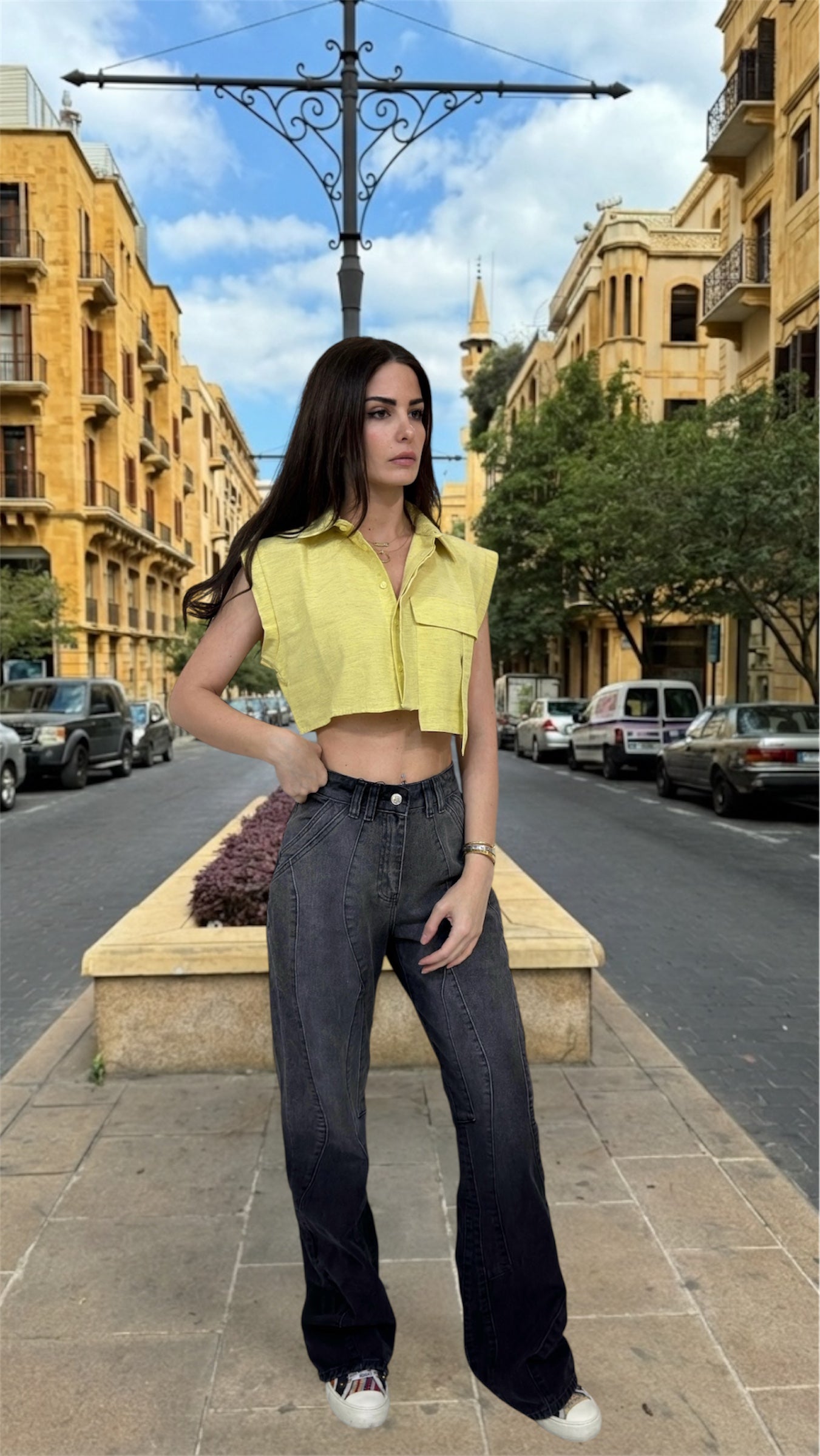 Woman in yellow top and black jeans standing on a street with buildings and cars in the background