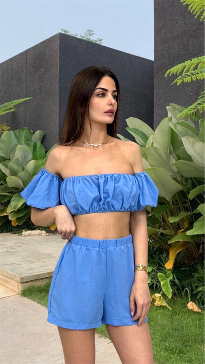 Woman wearing a blue off-shoulder top and shorts in an outdoor setting with plants and a building.