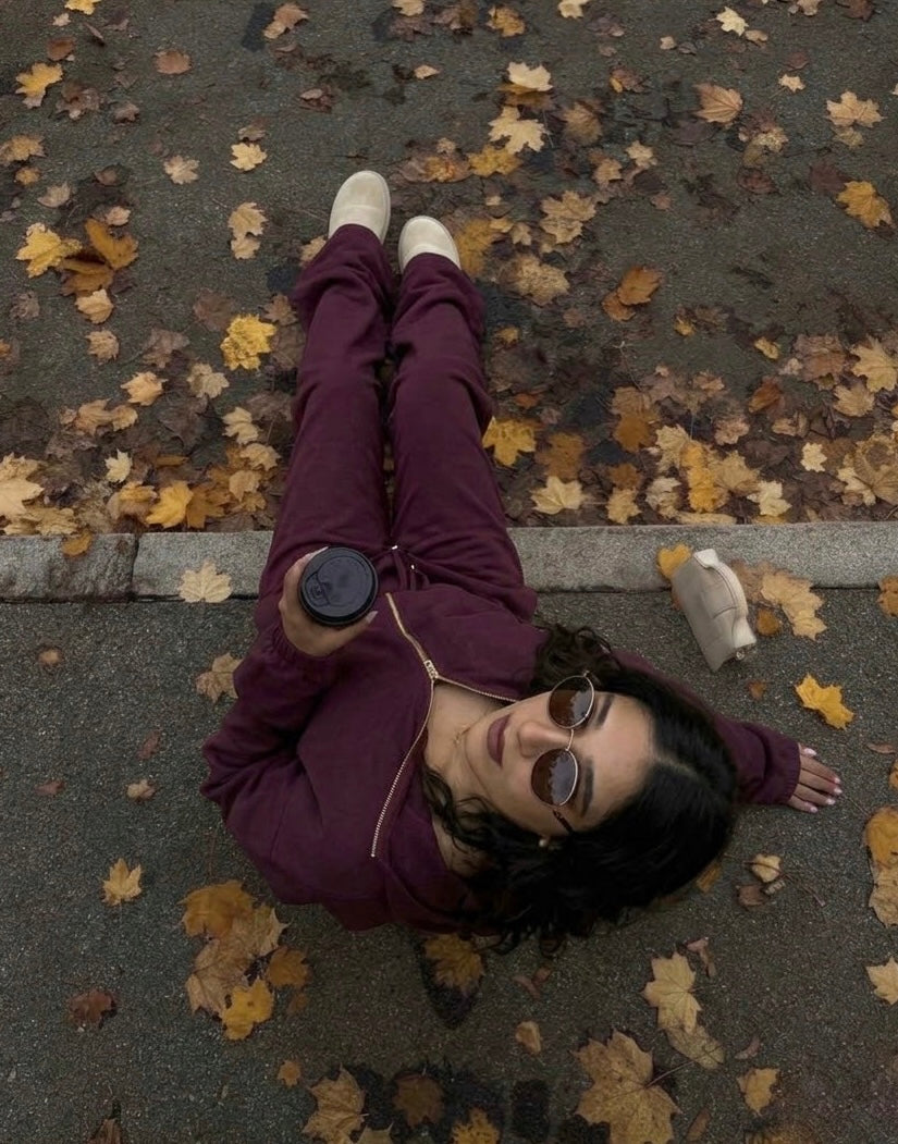 A woman lying on asphalt covered with fallen leaves, wearing a dark purple zip-up hoodie and matching sweatpants with white sneakers. She is holding a black coffee cup.